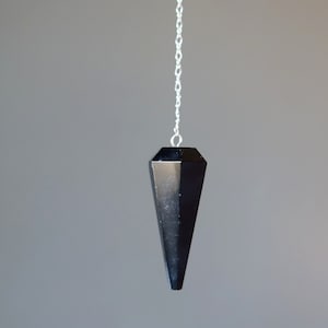 May include: A black crystal pendulum with a silver chain. The pendulum is shaped like a pointed cone.