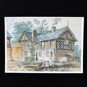 May include: A vintage print depicting a historic building with a timber-framed facade, stone base, and a slate roof. The scene includes trees, figures, and a smoking chimney, suggesting a rural setting. The artwork is framed with a white border.