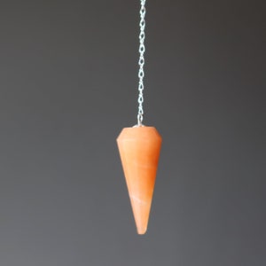 May include: An orange crystal pendulum suspended by a silver chain. The crystal is faceted with a pointed end. The background is a solid, muted gray.