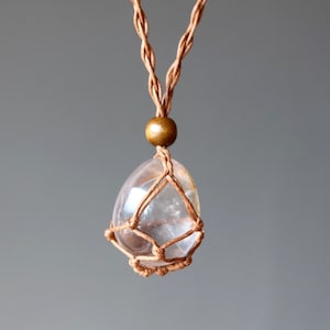 Puede incluir: Un colgante de cristal de cuarzo transparente cuelga de un collar de cordón de macramé marrón. El cristal está envuelto en un diseño de macramé anudado y tiene una cuenta de madera marrón en la parte superior.