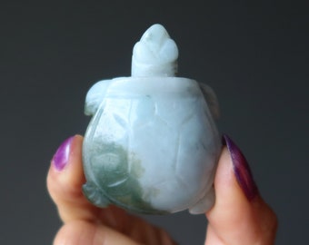 May include: A small jade turtle figurine with a light green and white color scheme. The turtle is carved in a detailed and realistic style, with a smooth, polished finish.