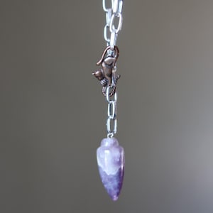May include: A silver chain with a copper cat charm and a purple and white amethyst pendulum. The pendulum is teardrop-shaped. The chain is hanging against a blurred gray background.