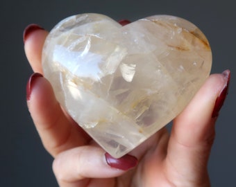 May include: A clear quartz heart with golden inclusions. The heart is polished and smooth, and it is held in a hand.