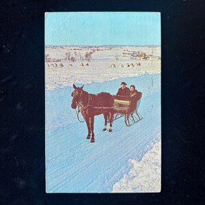 Peut inclure: Carte postale vintage représentant une calèche tirée par un cheval sur une route enneigée. Le cheval brun est attelé à un traîneau transportant deux personnes. L'arrière-plan montre un paysage enneigé sous un ciel bleu.