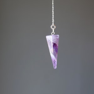 May include: A purple amethyst crystal pendulum on a silver chain. The crystal is faceted and has a pointed tip.