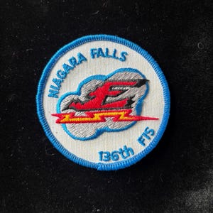 May include: Embroidered patch with a white background and a blue border. The patch features the text "NIAGARA FALLS" in blue, and "136th FIS" in blue. A red and yellow lightning bolt design is in the center.