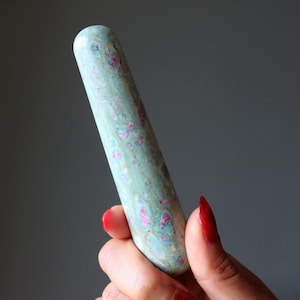 Puede incluir: Una varita de piedra pulida de forma cilíndrica, con un color base verde claro liso y motas irregulares de color magenta y rosa intenso dispersas por toda la superficie. La piedra tiene un extremo redondeado y una forma alargada y recta.