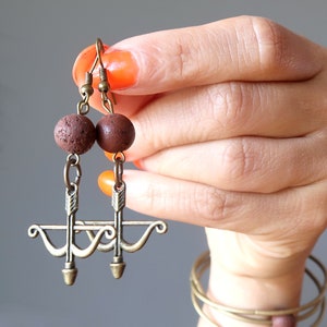 Puede incluir: Un par de pendientes marrones y dorados con un pequeño diseño de arco y flecha y cuentas de piedra marrón.