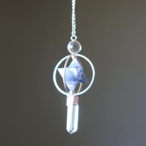 May include: A silver pendant featuring a clear crystal point and a blue star-shaped crystal. The pendant includes a clear crystal bead, a wire-wrapped design, and a silver chain. The design is suspended against a neutral background.