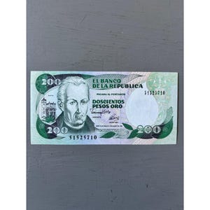 May include: A green and white 200 Colombian pesos banknote. The bill features a portrait of a man on the left, the text "EL BANCO DE LA REPUBLICA" at the top, and the denomination "DOSCIENTOS PESOS ORO". The serial number is 51525710.