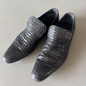 Pode incluir: Um par de sapatos pretos de couro sem cadarços. Os sapatos têm um padrão texturizado, semelhante ao de um réptil, e uma biqueira arredondada. Os sapatos são exibidos em uma superfície cinza claro. Os sapatos parecem não usados.