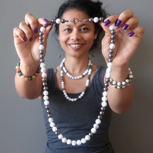 Puede incluir: Un collar largo con cuentas blancas y grises oscuras alternadas. El collar lo sostiene una persona que lleva una camiseta gris sin mangas.