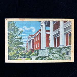 May include: Vintage postcard depicting Columbia College in Columbia, South Carolina. The image shows a red brick building with white columns and a green tree on the left. The sky is blue with some clouds. The postcard has the text "C-105 COLUMBIA COLLEGE, COLUMBIA, S. C."