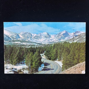 Puede incluir: Una postal vintage con un paisaje montañoso. La imagen muestra montañas cubiertas de nieve bajo un cielo azul, una carretera sinuosa y un denso bosque de árboles de hoja perenne. La postal tiene un aspecto ligeramente envejecido.