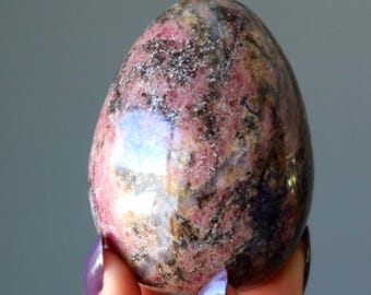 May include: A polished, egg-shaped stone featuring a mottled pattern of deep pink, black, and white mineral inclusions. The surface is smooth and reflective, showcasing a natural, variegated texture with swirling colors throughout the entire object.