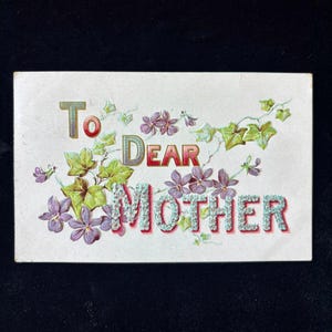 May include: A vintage postcard with the words "To Dear Mother" in red, green, and blue lettering. The card is decorated with purple flowers and green leaves, set against a white background.