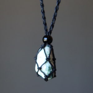 May include: A green and white agate stone pendant with black macrame knotting and a black bead. The pendant is hanging from a black cord necklace.