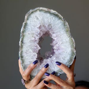 Può includere: Una grande geode di ametista di forma ovale con un centro cavo riempito di punte di cristallo traslucide color viola chiaro. Il bordo esterno della geode presenta una crosta minerale a strati e bande nei toni del grigio, bianco e verde pallido.