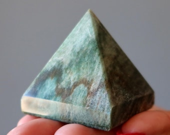 May include: A pyramid-shaped stone sculpture featuring a polished surface with a marbled pattern in shades of forest green, teal, and dark brown. The object has four triangular faces meeting at a sharp apex and a square base with smooth edges.