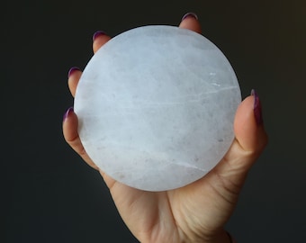 May include: A white selenite charging plate, a smooth, round disc of crystal, held in a hand.