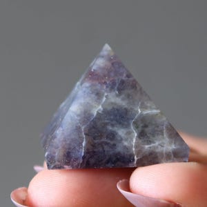 Peut inclure: Une petite sculpture en pierre en forme de pyramide avec une surface polie aux motifs marbrés violet foncé, violet et gris. La pierre présente des fractures internes naturelles et des veines sur toute sa structure, créant une texture unique.