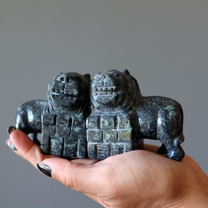 May include: Two dark green stone lion sculptures with detailed carvings. The lions are side-by-side, with a block-like structure between them. The sculptures are held in a hand against a grey background.