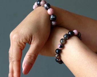 Puede incluir: Dos pulseras hechas con cuentas de piedras preciosas rosas y negras. Las pulseras se llevan en las muñecas de una persona.