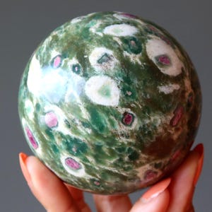 May include: A polished spherical stone featuring a mottled surface of deep forest green, interspersed with creamy white circular patches and small, distinct magenta-colored inclusions throughout the entire rounded mineral form.