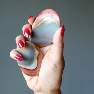Puede incluir: Dos piedras lisas de forma ovalada con un patrón en capas de gris, blanco y rojo. Las piedras se sostienen en una mano con esmalte de uñas rojo.