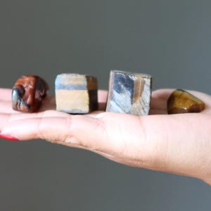May include: Four polished gemstones in shades of brown and gold are displayed. The stones vary in shape, including a rounded, tumbled stone and three geometric shapes. The gemstones are held in a hand against a neutral background.