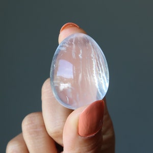 Puede incluir: Un cristal de selenita transparente, translúcido, de forma ovalada con estrías visibles. El cristal está siendo sostenido por una mano con esmalte de uñas rosa claro.