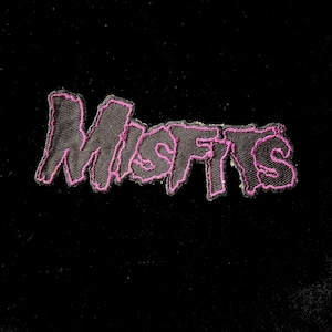 Op de afbeelding: Een zwarte patch met het woord "Misfits" geborduurd in een gestileerd lettertype met een roze omtrek. De patch is gemaakt van een textuurstof en is waarschijnlijk ontworpen om op kleding of accessoires te worden genaaid.