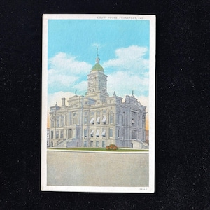 May include: Vintage postcard featuring the Courthouse in Frankfort, Indiana. The building is a large, ornate structure with a clock tower and green dome, set against a blue sky with fluffy clouds. The postcard has a white border and the text "COURT HOUSE, FRANKFORT, IND".