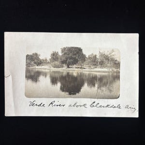 May include: A vintage black and white photograph of the Verde River above Clarkdale, Arizona. The image shows a calm river reflecting trees on the bank, with the handwritten text 'Verde River above Clarkdale Ariz.' visible below the photo frame.