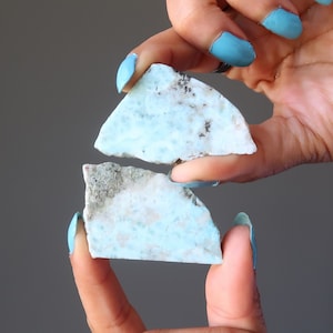 May include: Two pieces of blue and white ocean jasper, a type of chalcedony, are held in a hand with blue nail polish. The stone has a unique pattern of white and blue.