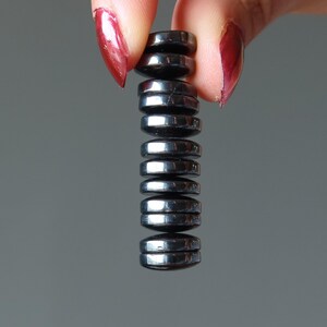 May include: A stack of small, shiny, dark gray magnetic hematite discs. The discs are held between two fingers with red nail polish against a gray background. The discs are stacked vertically.
