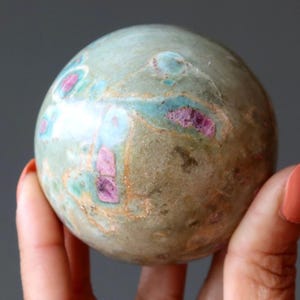 May include: A polished spherical stone featuring a muted sage green base color with prominent inclusions of vibrant magenta and streaks of light blue. The surface displays a smooth, rounded texture with natural mineral patterns throughout the sphere.