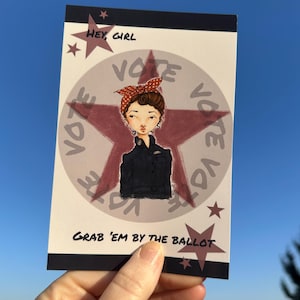 May include: A postcard featuring a stylized illustration of a woman with a red and brown polka dot headscarf, set against a red star and the word "VOTE". The text "HEY, GIRL" and "GRAB 'EM BY THE BALLOT" are also included.