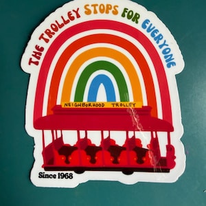 Pu&ograve; includere: Un adesivo raffigurante un tram rosso con passeggeri, ambientato su un arcobaleno. Il testo recita "The Trolley Stops for Everyone" e "Neighborhood Trolley" con "Since 1968" in basso. L'adesivo ha un bordo bianco.