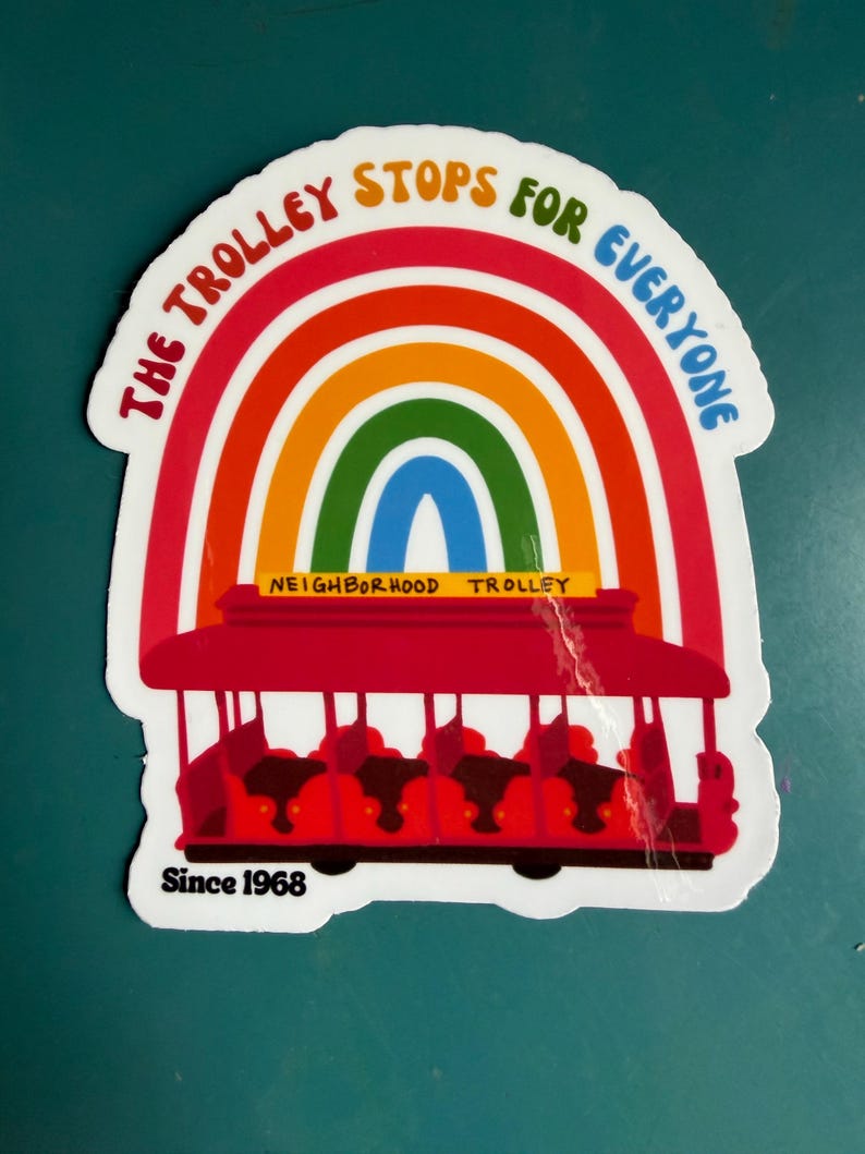 Pu&ograve; includere: Un adesivo raffigurante un tram rosso con passeggeri, ambientato su un arcobaleno. Il testo recita "The Trolley Stops for Everyone" e "Neighborhood Trolley" con "Since 1968" in basso. L'adesivo ha un bordo bianco.