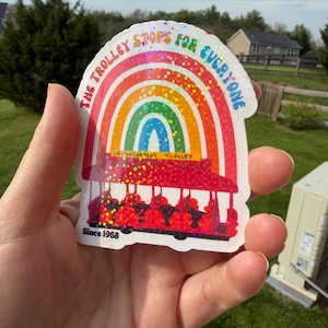 May include: A vibrant sticker with a rainbow design and the words "THE TROLLEY STOPS FOR EVERYONE." A red trolley car is depicted below the rainbow. The sticker also includes the text "NEIGHBORHOOD TROLLEY" and "Since 1968."