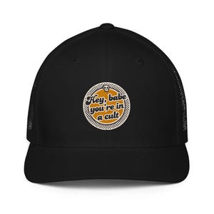 May include: A black trucker hat featuring a circular patch on the front. The patch has a mustard yellow center with the black text 'Hey, babe you're in a cult' and a small skull icon at the top, surrounded by a black and white checkered border.