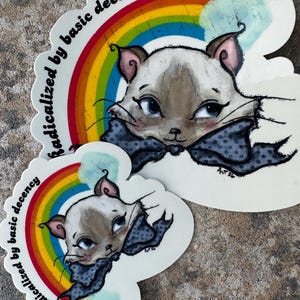 K&ouml;nnte beinhalten: Zwei Aufkleber mit einer Katze mit blauen Augen und einer gepunkteten Fliege, vor einem Regenbogen. Der Text "radicalized by basic decency" ist um den Regenbogen herum angeordnet. Die Katze ist hellbraun und wei&szlig;.