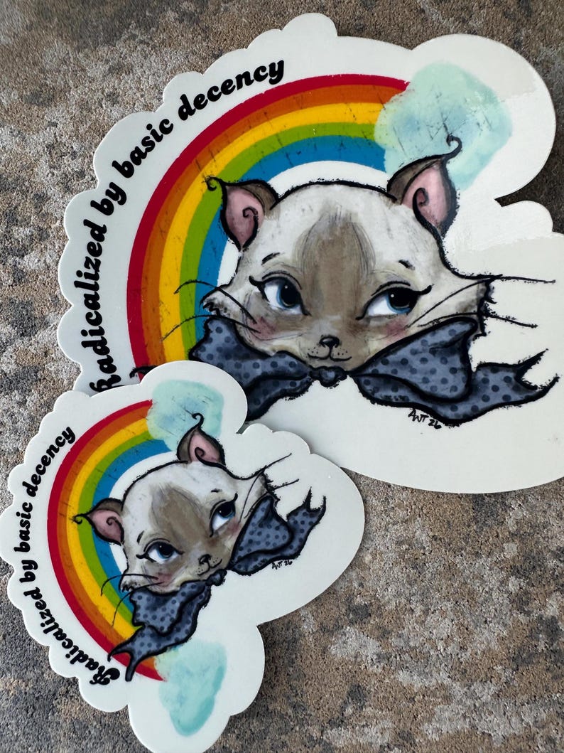 K&ouml;nnte beinhalten: Zwei Aufkleber mit einer Katze mit blauen Augen und einer gepunkteten Fliege, vor einem Regenbogen. Der Text "radicalized by basic decency" ist um den Regenbogen herum angeordnet. Die Katze ist hellbraun und wei&szlig;.