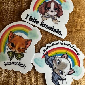Könnte beinhalten: Drei gestanzte Aufkleber mit Zeichentricktieren und Regenbogen. Einer zeigt einen Hund mit dem Text "I bite fascists." Ein anderer zeigt eine Katze mit "Jesus was Antifa." Der dritte zeigt eine Katze mit "Radicalized by basic decency."