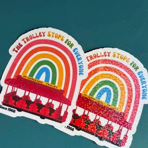 May include: Two stickers with a red trolley design, a rainbow arch, and the text "The Trolley Stops For Everyone." The stickers have a white border and the words "Neighborhood Trolley" and "Since 1968."