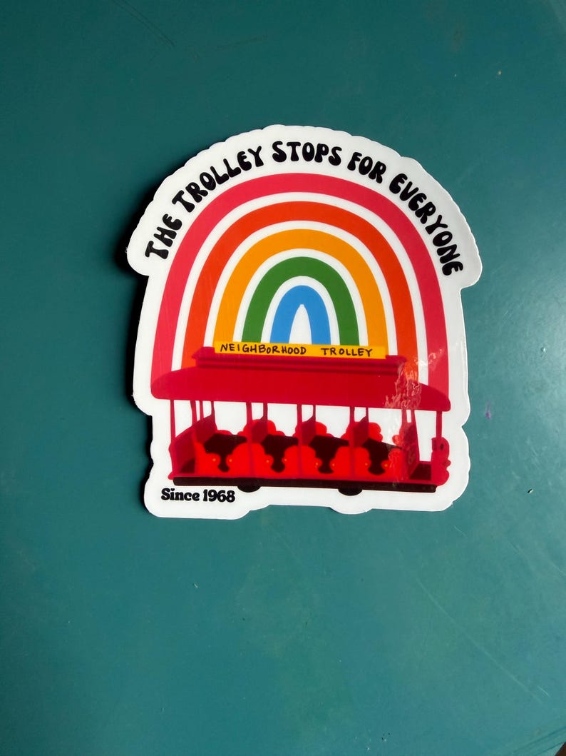 Pu&ograve; includere: Un adesivo raffigurante un tram rosso con un arcobaleno sopra. Il testo "THE TROLLEY STOPS FOR EVERYONE" &egrave; arcuato sopra l'arcobaleno. Le parole "NEIGHBORHOOD TROLLEY" sono sotto, e "Since 1968" in basso.