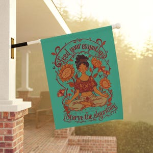 May include: A teal garden flag featuring a central illustration of a person meditating in a lotus position, surrounded by sunflowers. The text "Feed your empathy" is printed above and "Starve the algorithm" is printed below in dark red.