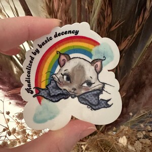K&ouml;nnte beinhalten: Ein Aufkleber mit einer Katze mit blauer Schleife, Regenbogen und dem Text "radicalized by basic decency". Die Katze ist grau und wei&szlig;. Der Aufkleber wird von einer Person gehalten.
