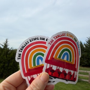 Pu&ograve; includere: Due adesivi con un design arcobaleno e la scritta "The Trolley Stops for Everyone". Gli adesivi presentano un tram rosso e un arco arcobaleno. &Egrave; visibile anche la parola "Neighborhood".